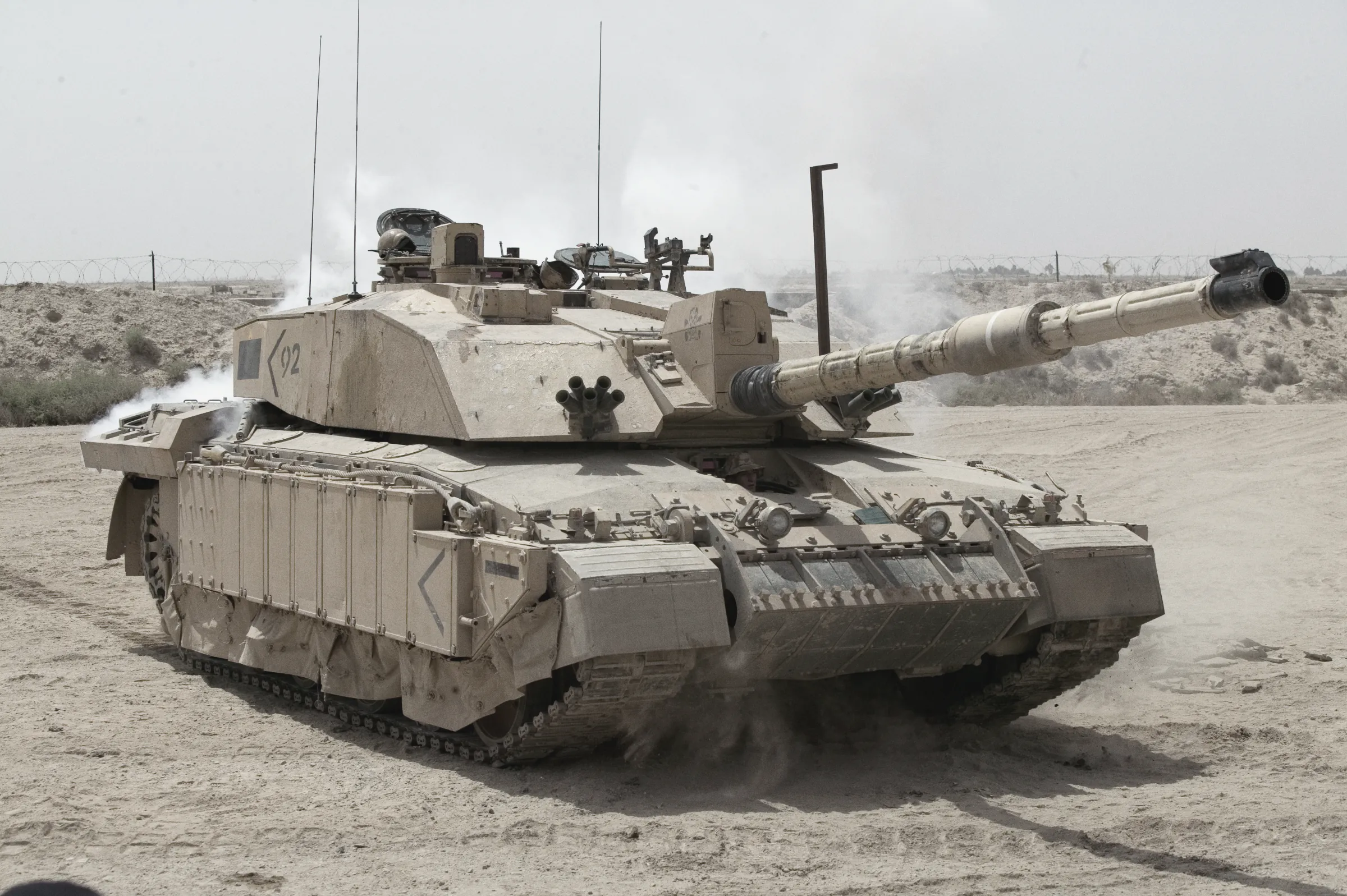 A British Challenger 2 tank firing on patrol in open desert outside Basra on 1 June 2004, smoke from its barrel and dust thrown up by its tracks, with barbed wire in the foreground.