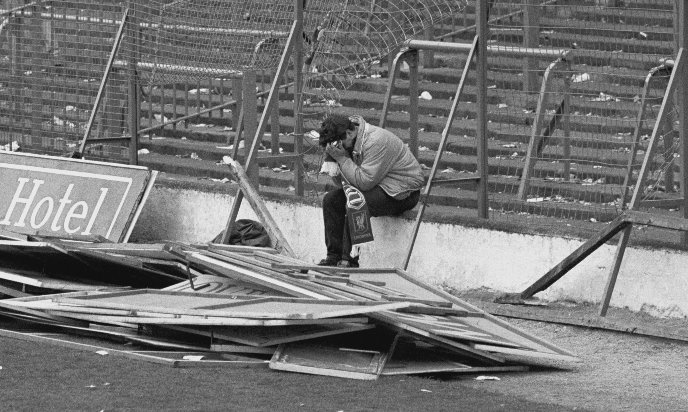 The empty Leppings Lane terrace at Hillsborough after the crush on 15 April 1989, behind its perimeter fence, with broken advertising boards piled on the pitch and a Liverpool supporter sitting alone on the wall, his face in his hands.