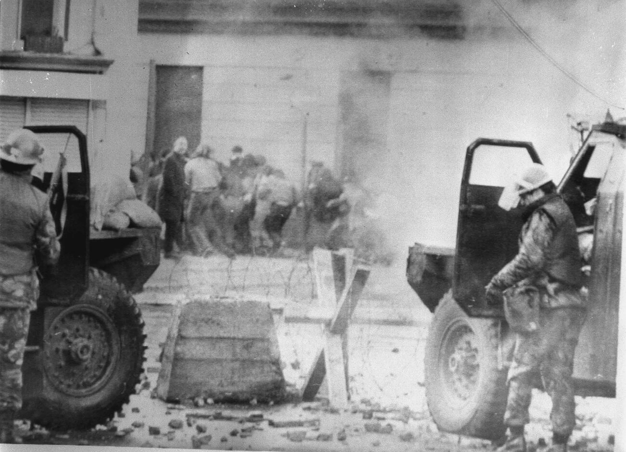 Two British soldiers in helmets shelter behind their armoured vehicles at a barbed-wire barricade in Derry on 30 January 1972, stones on the road, with a crowd half-hidden in CS gas beyond.