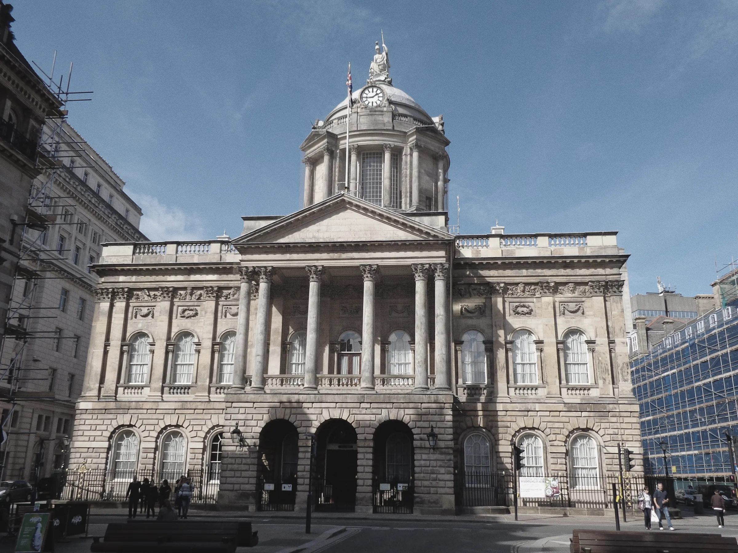 The front of Liverpool Town Hall in daylight under a blue sky: a columned portico above three arched entrances, with a clock and domed cupola behind, and a few pedestrians small in the street.