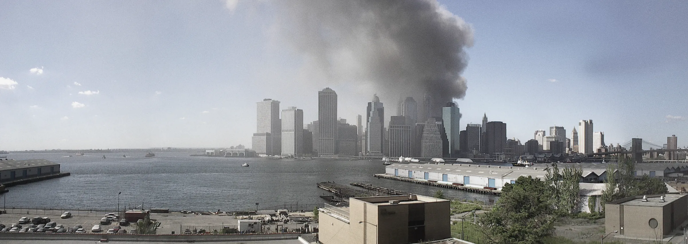 Lower Manhattan seen across the harbour from Brooklyn on the morning of 11 September 2001, both towers of the World Trade Center burning and a plume of smoke drifting across the sky.