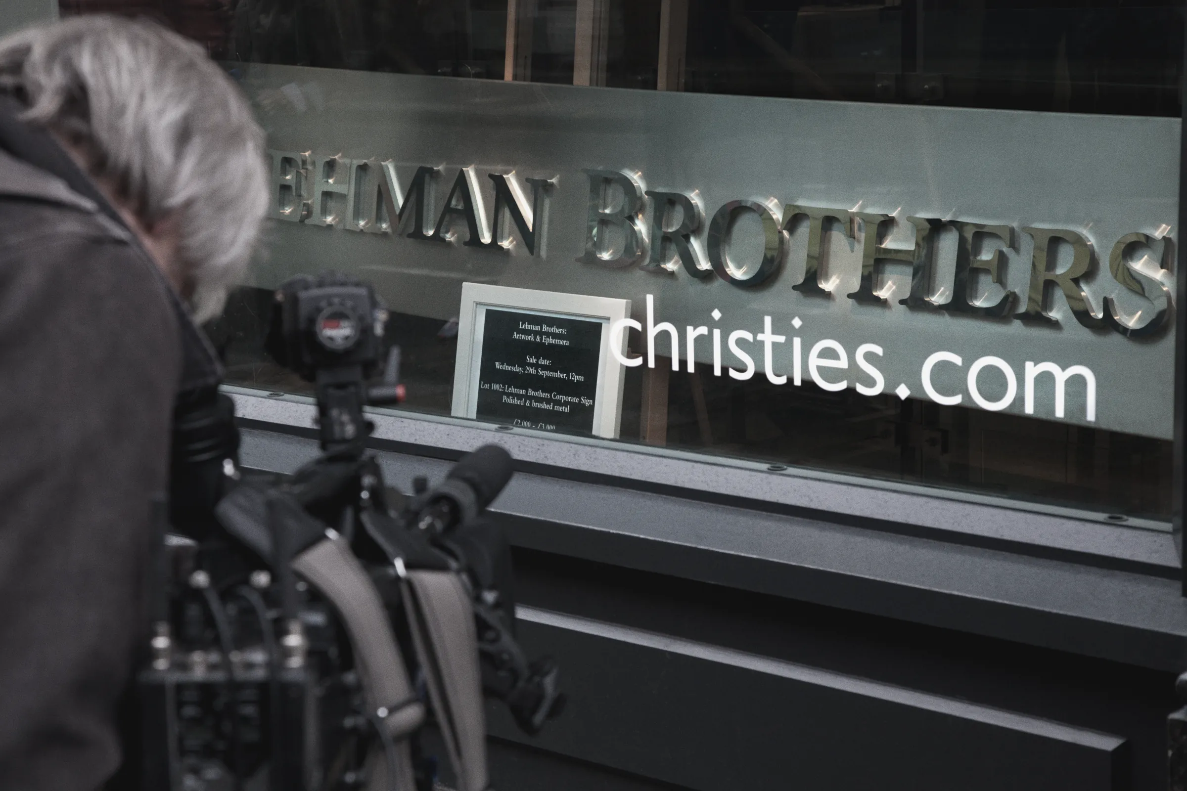A television camera crew filming Lehman Brothers' illuminated corporate sign in a Christie's shop window, London, beside a card advertising the sale of "Lehman Brothers: Artwork & Ephemera."