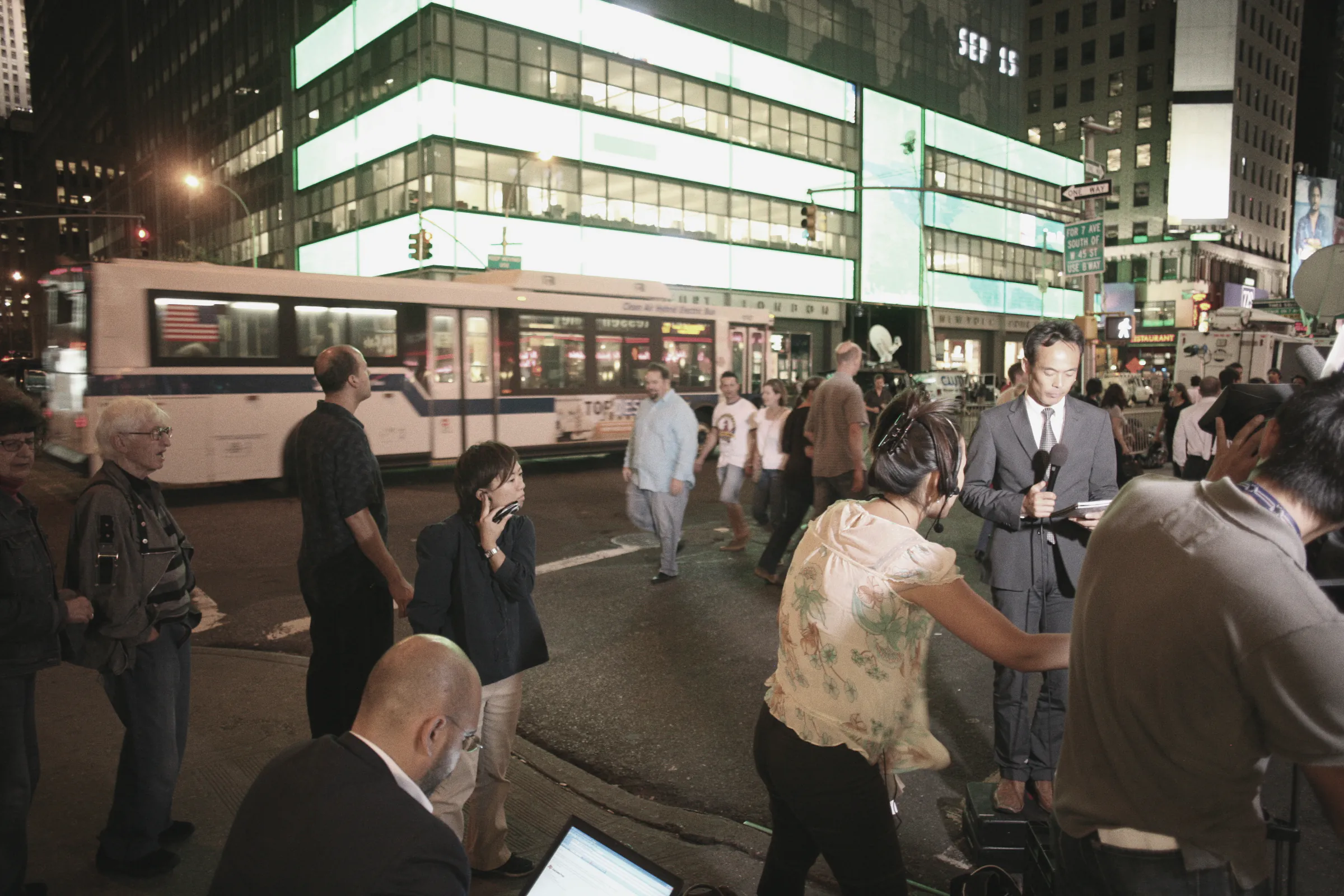 Television crews and onlookers on Seventh Avenue at night outside the lit headquarters of Lehman Brothers, its ticker reading "SEP 15", on the day the bank filed for bankruptcy.
