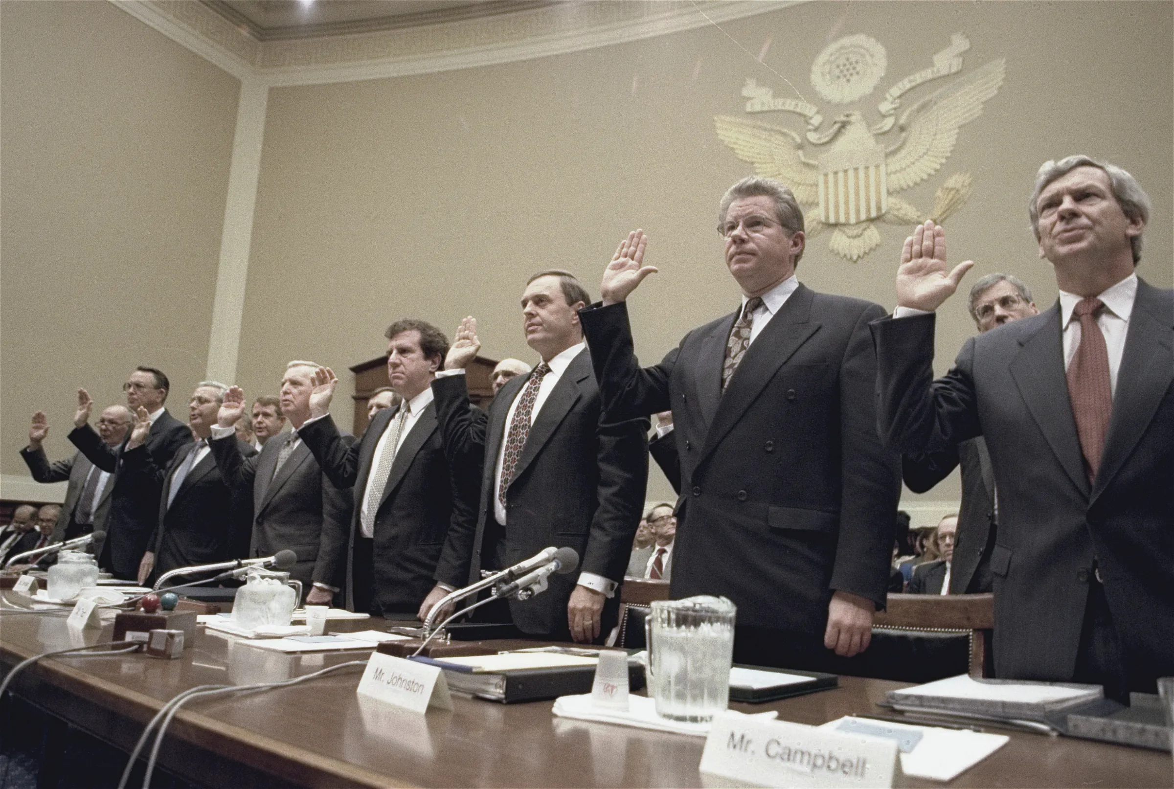The heads of seven tobacco companies stand with their right hands raised to be sworn in before a House subcommittee on 14 April 1994, Philip Morris's William Campbell nearest the camera behind his nameplate.